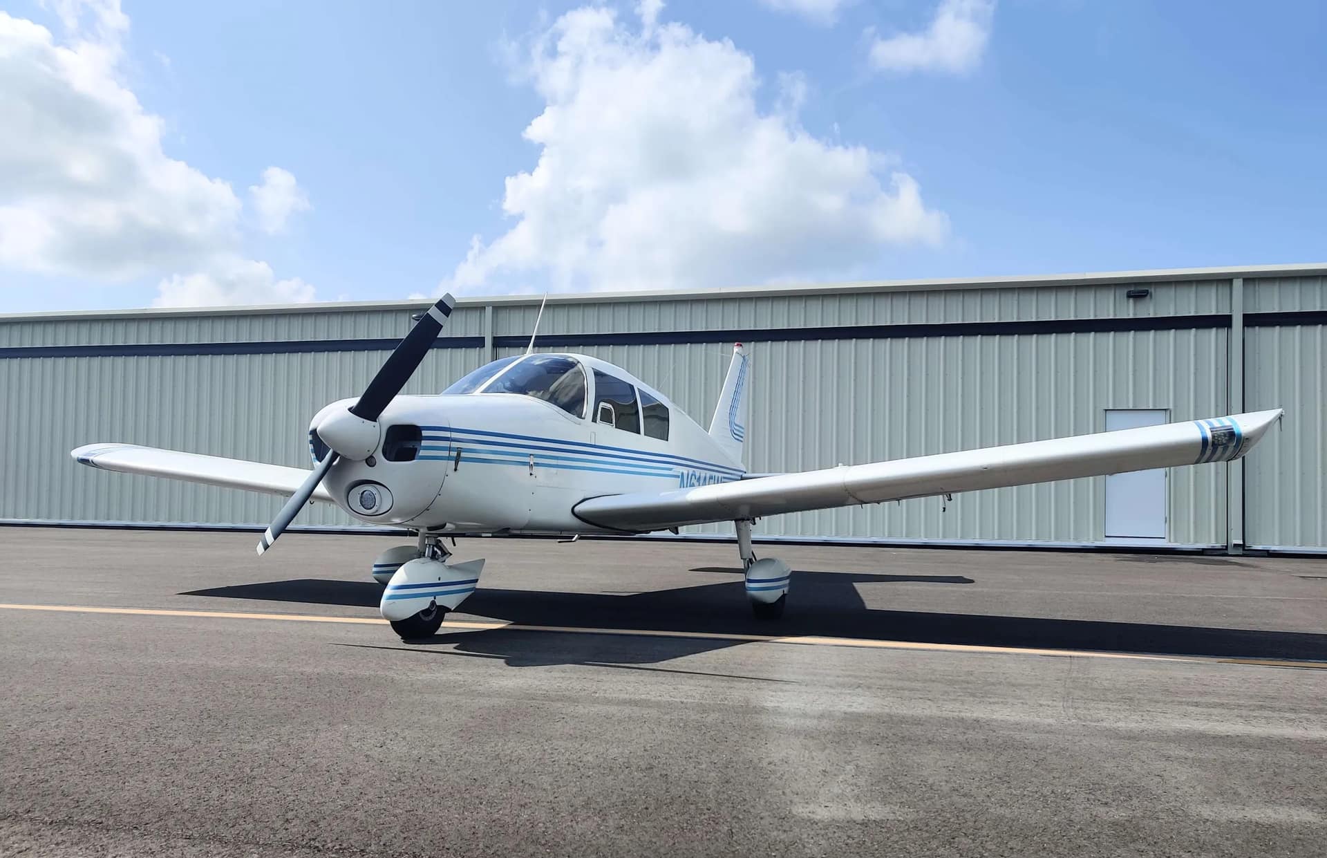 White-and-blue Piper Cherokee 140 parked on the ramp in front of a hangar under a blue Florida sky